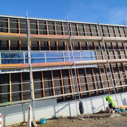 WOODEN - CON SPÓŁKA Z OGRANICZONĄ ODPOWIEDZIALNOŚCIĄ - Budowa domu szkieletowego: widok na elewację z rusztowaniem, widoczne drewniane belki konstrukcyjne i czarna membrana, pracownik w odblaskowej kamizelce na ziemi.