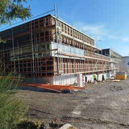 WOODEN - CON SPÓŁKA Z OGRANICZONĄ ODPOWIEDZIALNOŚCIĄ - Budowa domu szkieletowego z widocznymi rusztowaniami, drewnianą konstrukcją ścian i elementami izolacji, na tle błękitnego nieba.