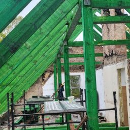 WOODEN - CON SPÓŁKA Z OGRANICZONĄ ODPOWIEDZIALNOŚCIĄ - Malowana na zielono drewniana konstrukcja więźby dachowej w trakcie montażu, widoczne rusztowania i fragmenty murowanego komina