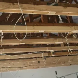 Wnętrze w trakcie prac wykończeniowych: widok na drewniany szkielet dachu z widocznymi przewodami elektrycznymi, częściowo zakryty płytami kartonowo-gipsowymi.