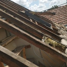 WOODEN - CON SPÓŁKA Z OGRANICZONĄ ODPOWIEDZIALNOŚCIĄ - Demontaż starego dachu z widoczną więźbą dachową, fragmentami izolacji i nowym oknem dachowym w trakcie montażu, na tle błękitnego nieba z chmurami.