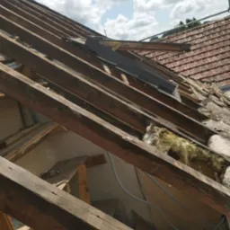 Demontaż starego dachu z widoczną więźbą dachową, fragmentami izolacji i nowym oknem dachowym w trakcie montażu, na tle błękitnego nieba z chmurami.