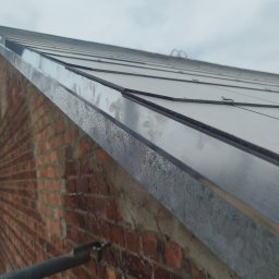 WOODEN - CON SPÓŁKA Z OGRANICZONĄ ODPOWIEDZIALNOŚCIĄ - Metalowy, płaski dach z widoczną krawędzią i ceglaną ścianą budynku. Ujęcie z dołu, podkreślające detale wykończenia i fakturę materiałów.