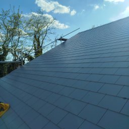 WOODEN - CON SPÓŁKA Z OGRANICZONĄ ODPOWIEDZIALNOŚCIĄ - Szary dach z dachówki bitumicznej, częściowo widoczne rusztowanie, błękitne niebo z chmurami w tle. Widok z dołu, podkreślający strukturę pokrycia.