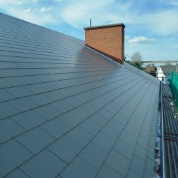 WOODEN - CON SPÓŁKA Z OGRANICZONĄ ODPOWIEDZIALNOŚCIĄ - Szary dach pokryty dachówką ceramiczną z ceglanym kominem w tle, widziany z góry. Widoczne rusztowania i fragmenty sąsiednich budynków.