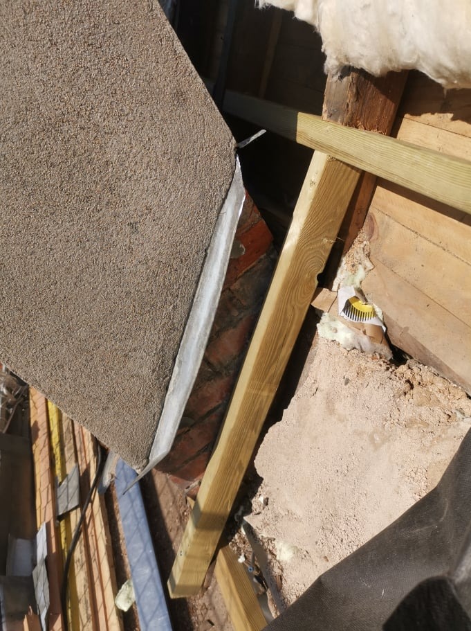 Widok z góry na fragment dachu w trakcie prac: połączenie starej ściany z cegły, nowej drewnianej konstrukcji i fragmentu pokrycia z papy, widoczne ocieplenie z wełny mineralnej.