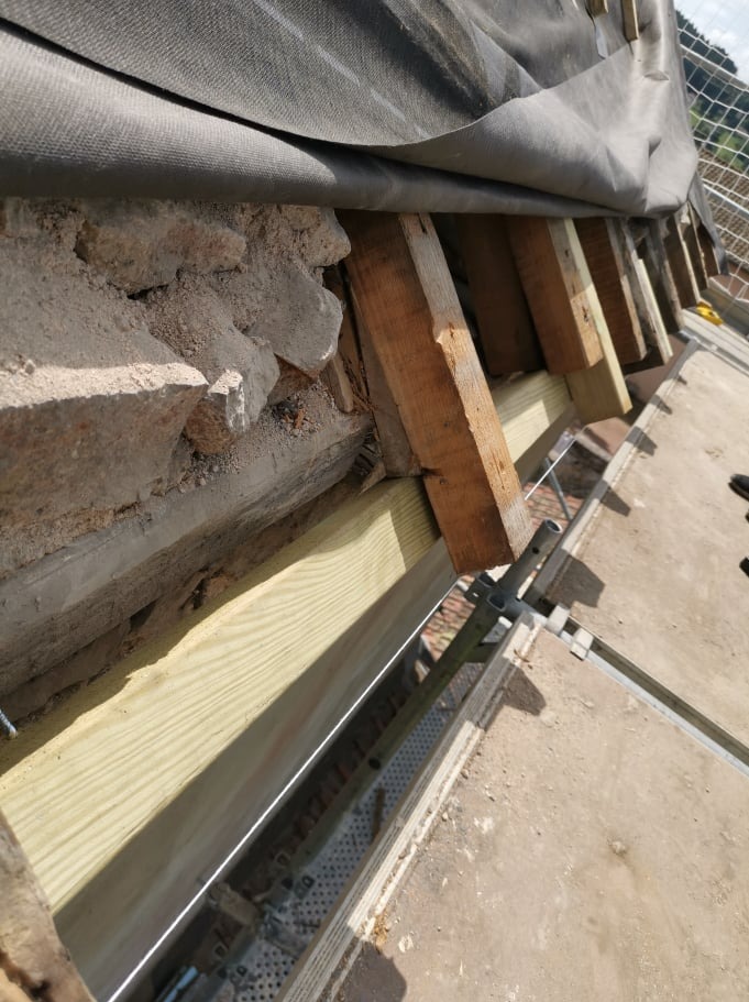 Fragment konstrukcji dachu z widoczną starą cegłą, nowymi drewnianymi belkami i szarą membraną dachową, widziany z bliska z rusztowania.