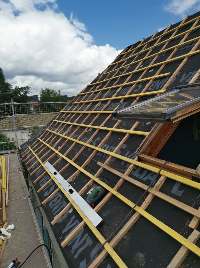 Montaż łat na dachu z oknem dachowym, widoczna poziomica i piła elektryczna na czarnej membranie dachowej.