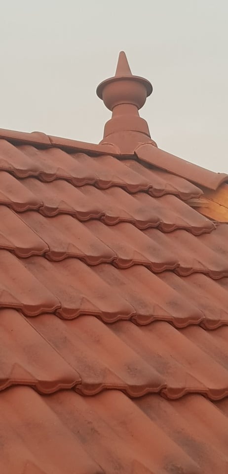 Czerwona dachówka ceramiczna z ozdobnym zwieńczeniem w kształcie stożka na szczycie dachu, widok z dołu na tle pochmurnego nieba.