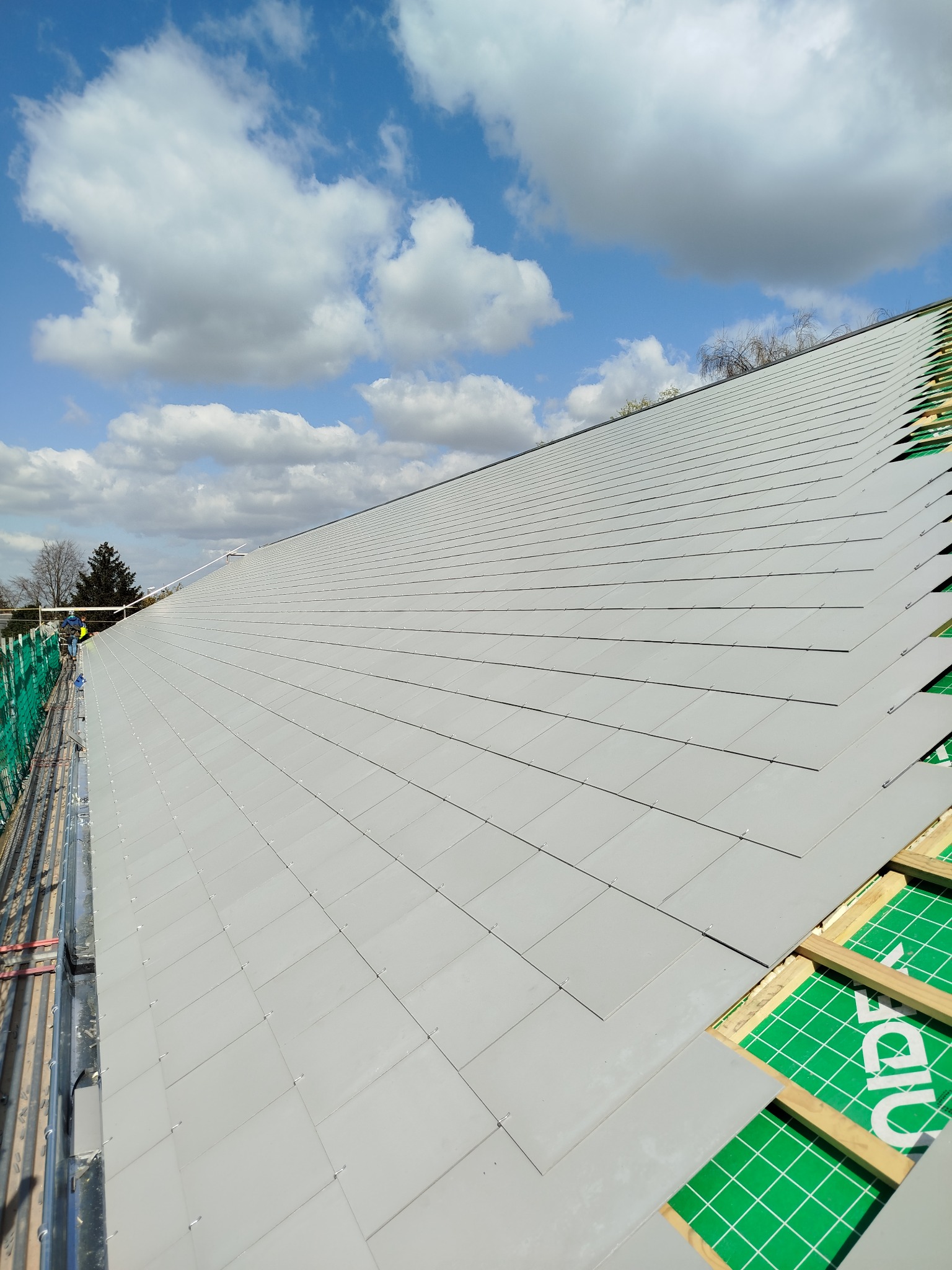 Układanie szarej dachówki na skośnym dachu. Widoczne membrany dachowe i fragment rusztowania. Błękitne niebo z chmurami w tle.