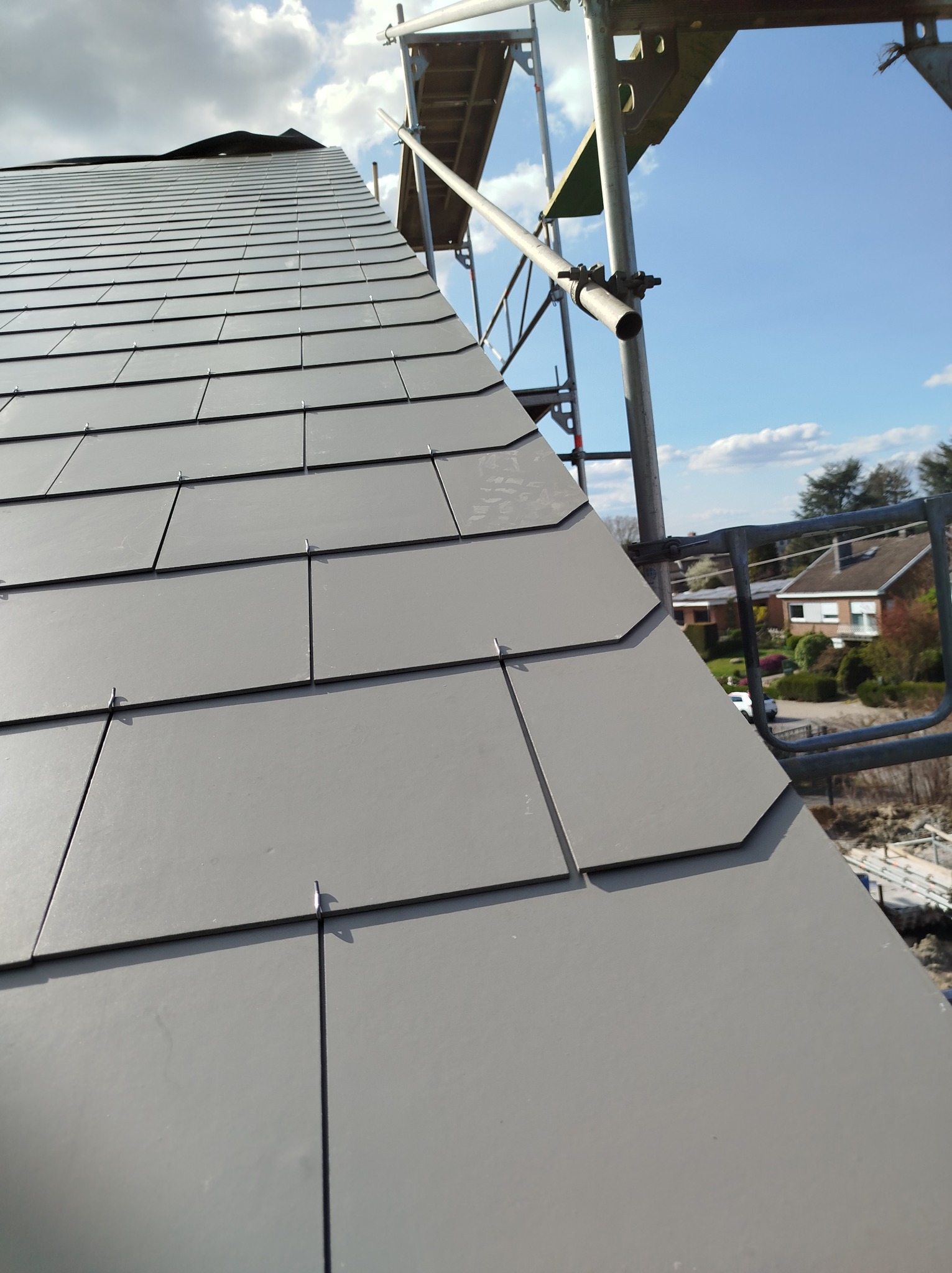 Szare, połaciowe krycie dachu z widocznymi elementami rusztowania na tle błękitnego nieba i zabudowań mieszkalnych. Widok z bliska.