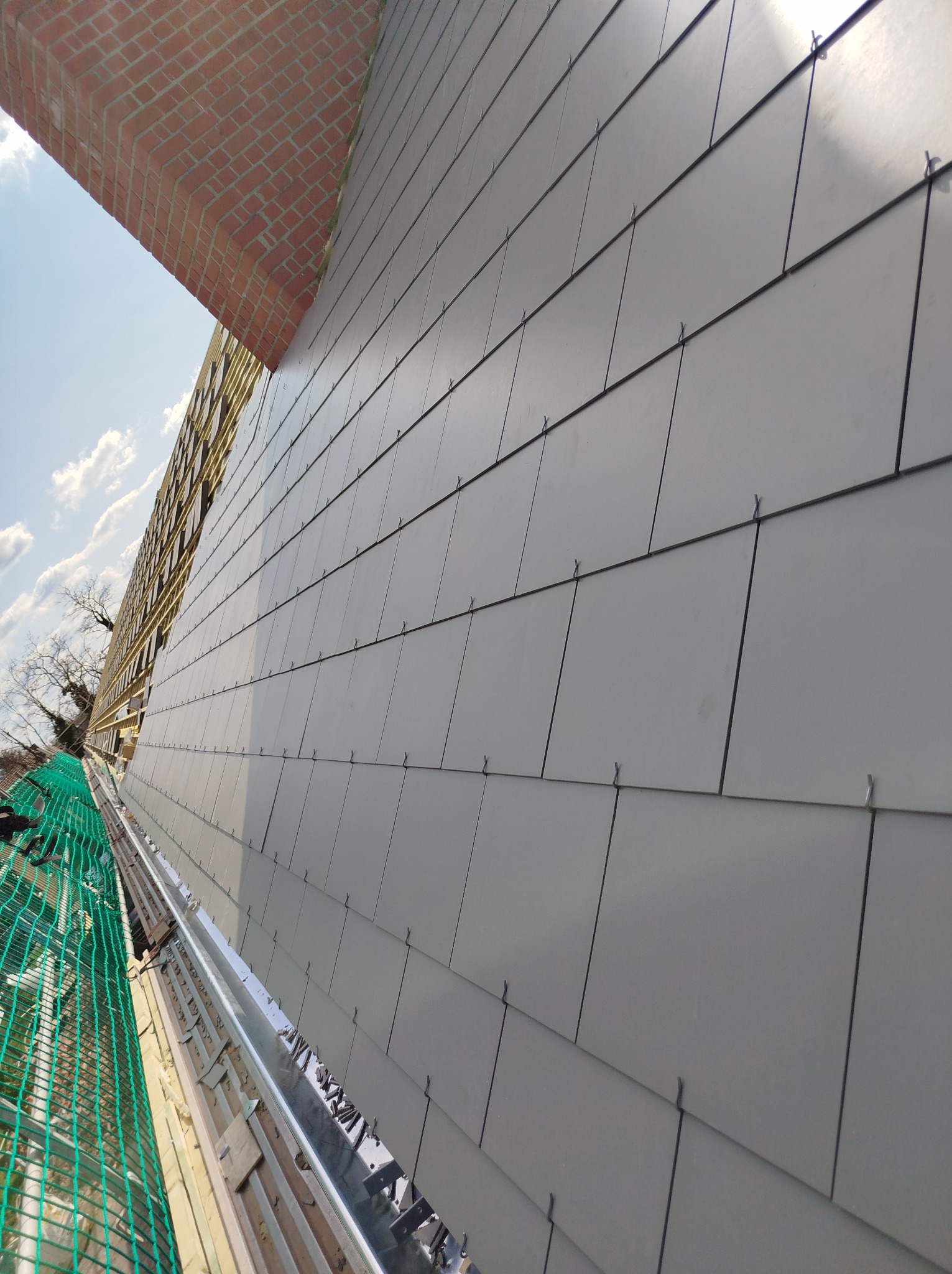 Szare panele dachowe ułożone na dachu, widoczne elementy konstrukcyjne i fragment komina z cegły. Ujęcie z dołu, podkreślające regularność wzoru paneli.