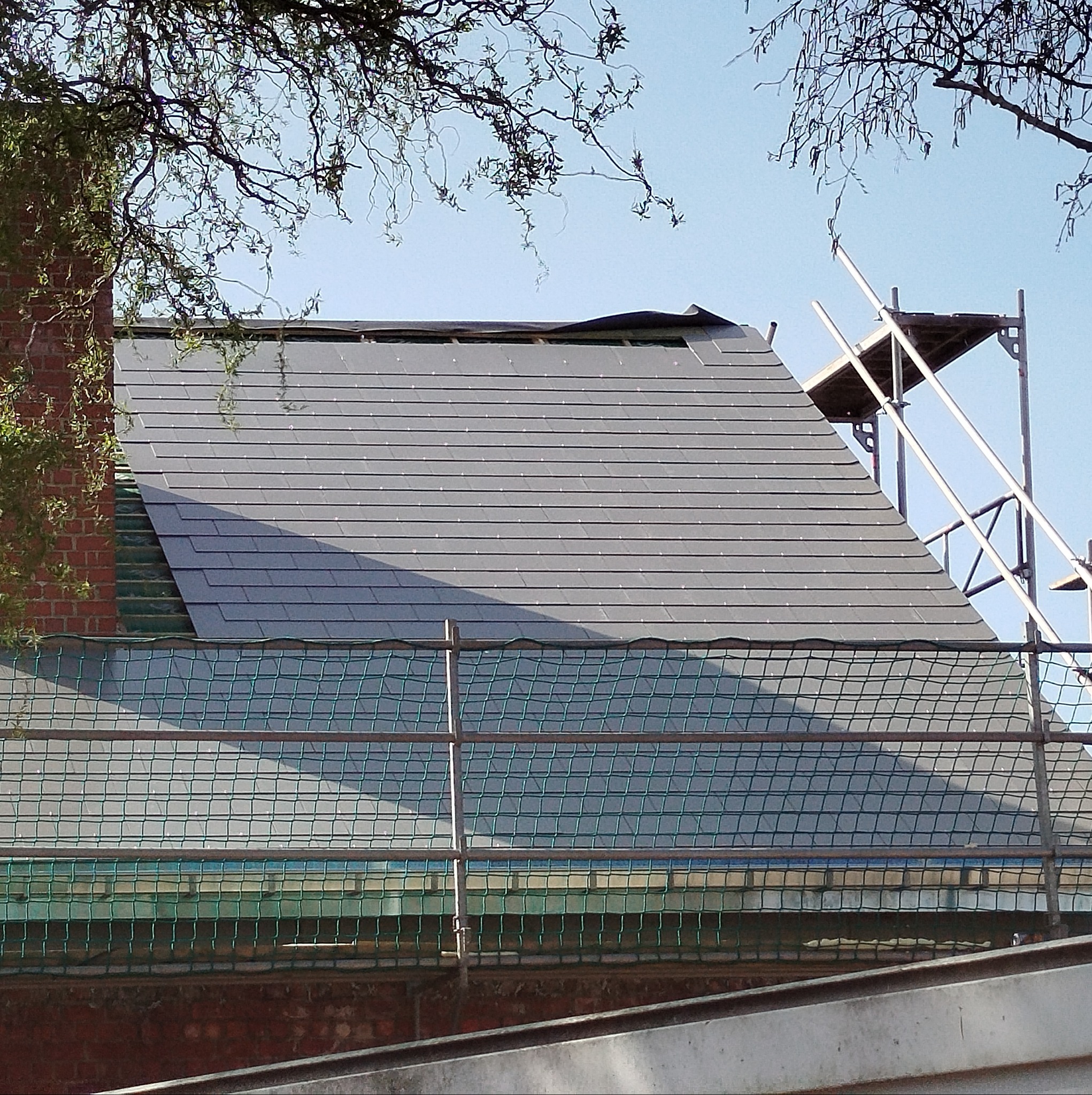 Krycie dachu szarą dachówką, widok z rusztowania, Strzelce Krajeńskie. Częściowo ukończone prace dekarskie, widoczny komin i korona drzewa.