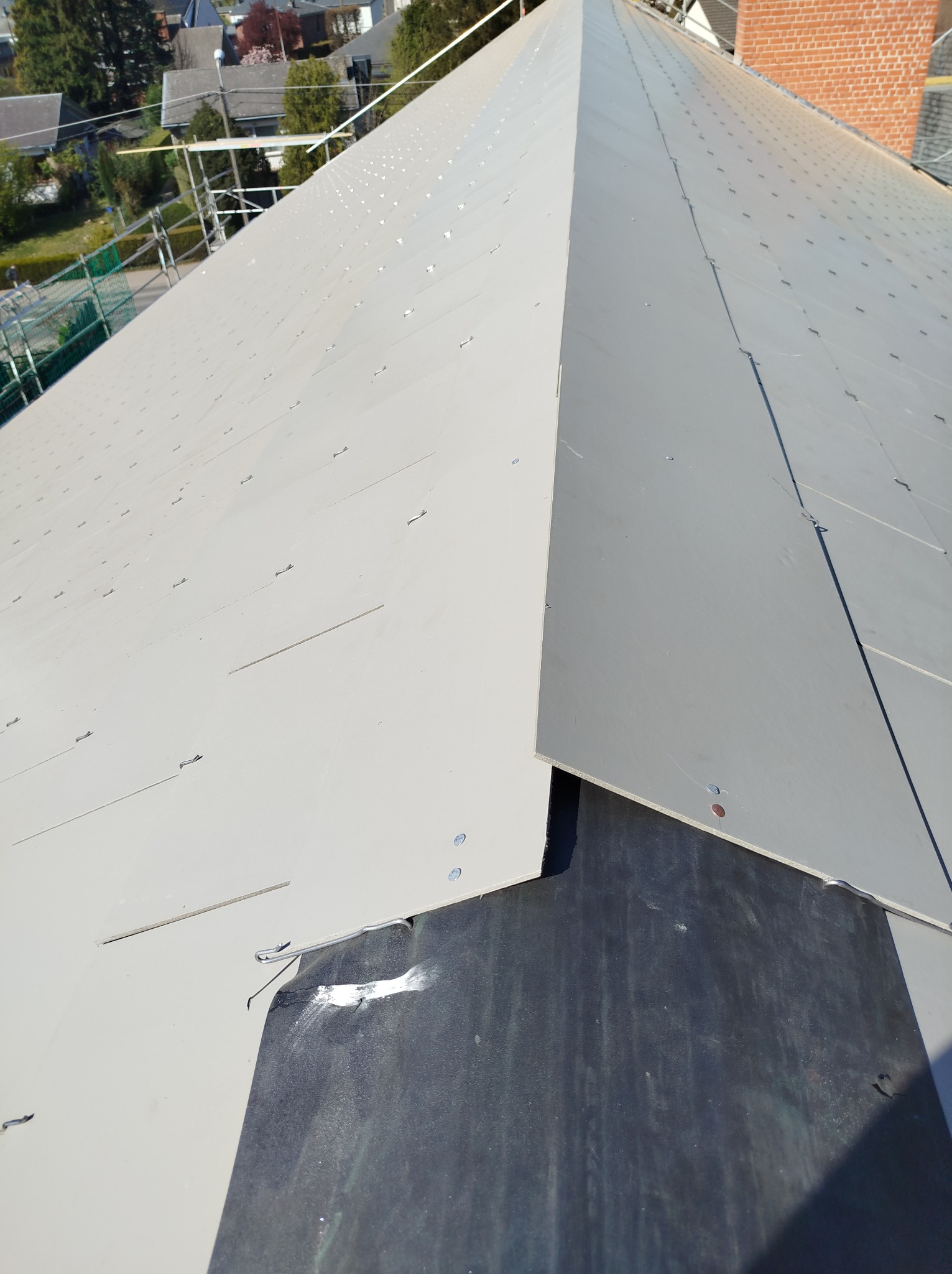Szary dach kryty panelami na wkręty, widok z góry, zarys komina w tle. Widoczny fragment czarnego podłoża pod panelami i elementy rusztowania.