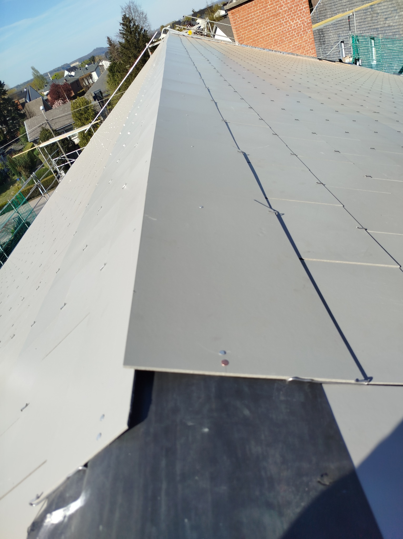 Nowe, szare pokrycie dachu z widocznymi mocowaniami, ułożone na czarnej membranie dachowej. W tle zabudowa mieszkalna i błękitne niebo. Kąt ujęcia z góry.