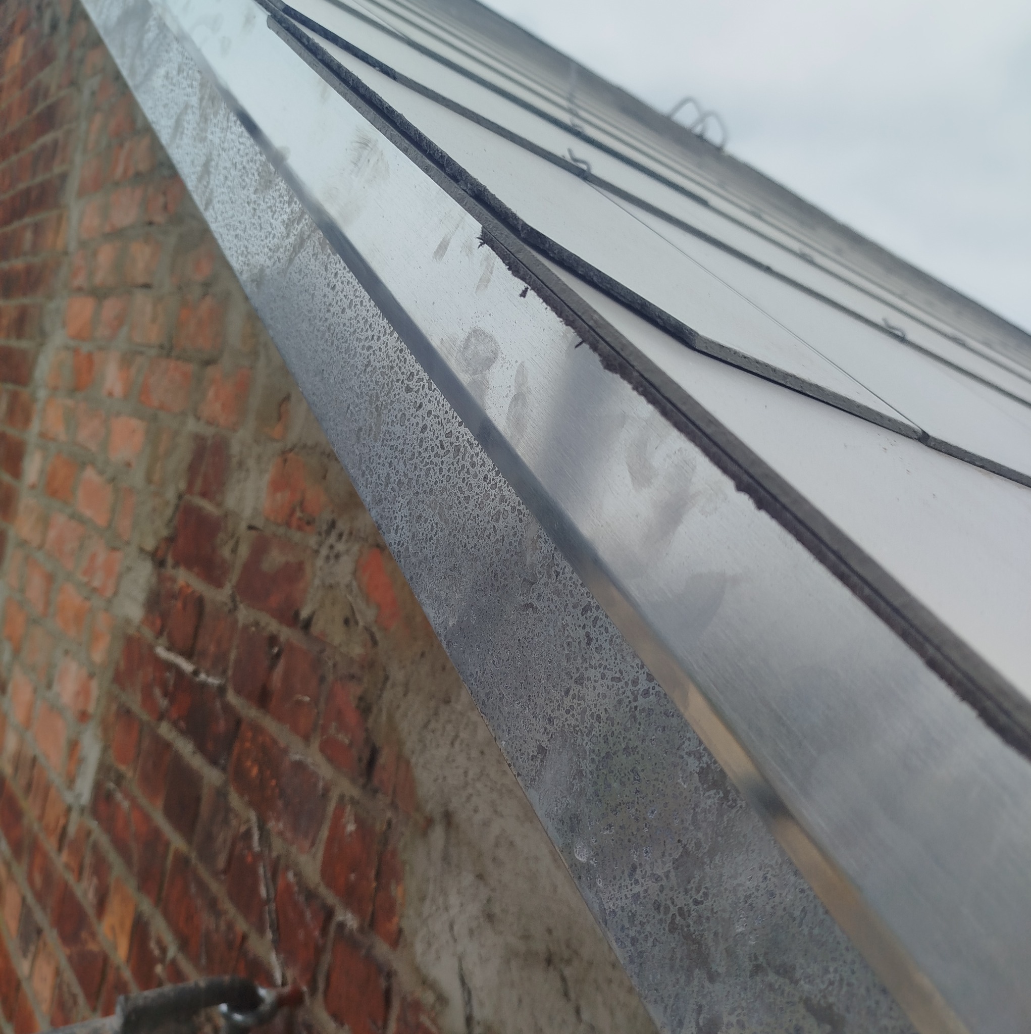 Fragment dachu z blachy trapezowej, widoczny z bliska, z detalami łączeń i fakturą materiału. W tle mur z czerwonej cegły i fragment nieba.