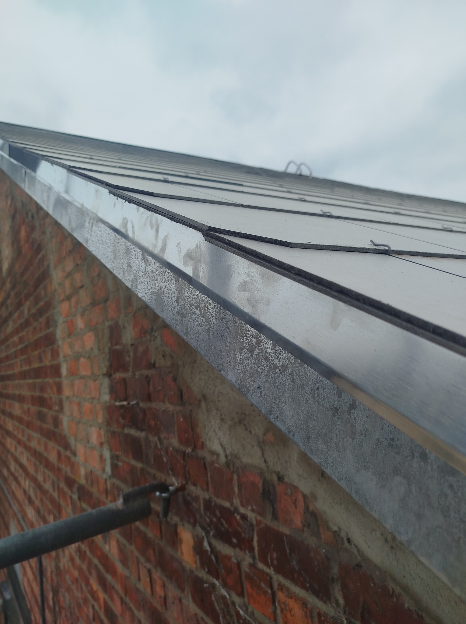 Metalowy, płaski dach z widoczną krawędzią i ceglaną ścianą budynku. Ujęcie z dołu, podkreślające detale wykończenia i fakturę materiałów.