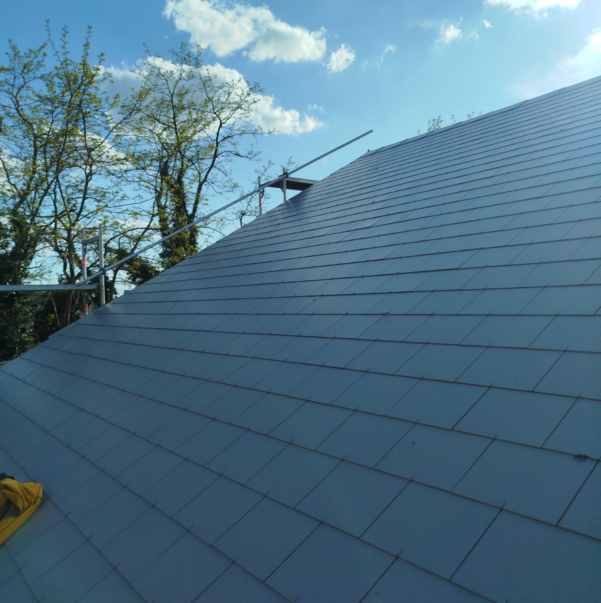 Szary dach z dachówki bitumicznej, częściowo widoczne rusztowanie, błękitne niebo z chmurami w tle. Widok z dołu, podkreślający strukturę pokrycia.