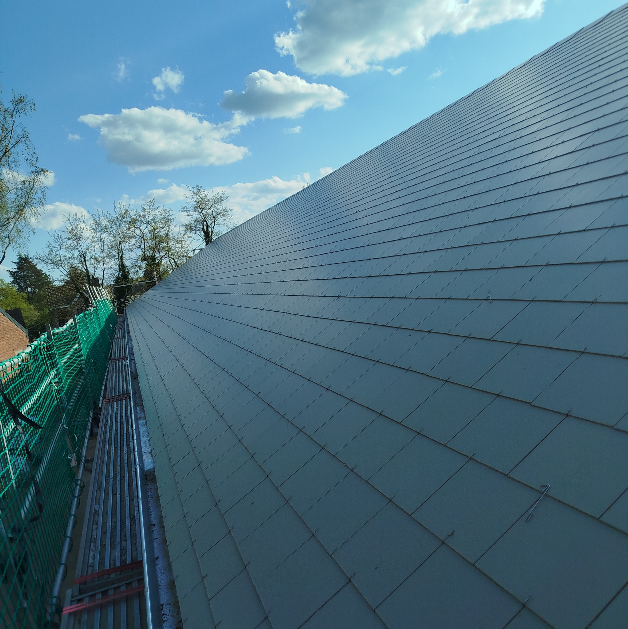 Niebiesko-szary dach pokryty dachówką bitumiczną widziany z rusztowania, z zieloną siatką zabezpieczającą i błękitnym niebem w tle.