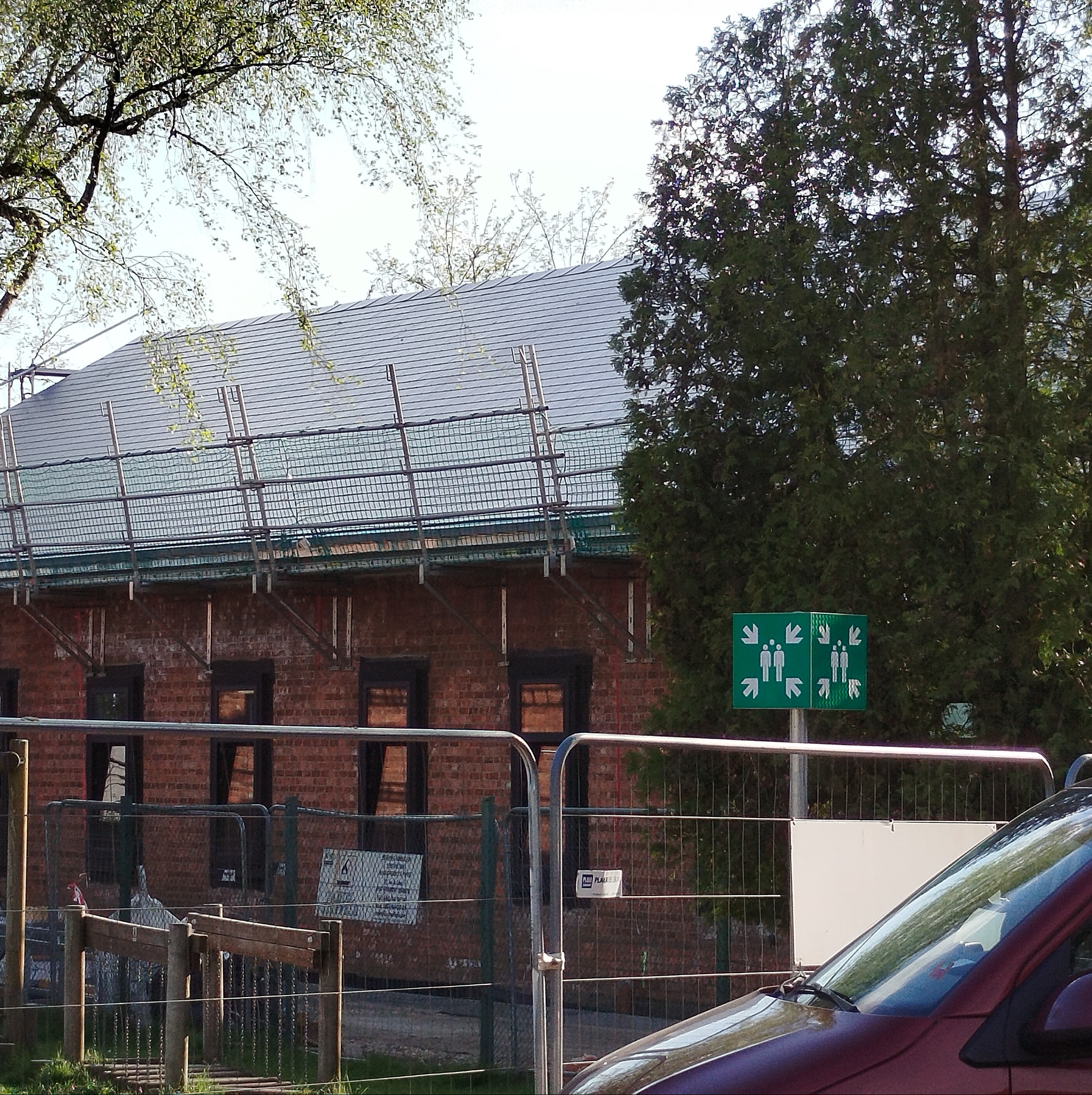 Ciemna dachówka na budynku z czerwonej cegły, zabezpieczona siatką rusztowaniową. Widoczny fragment ogrodzenia i zielona tablica informacyjna.