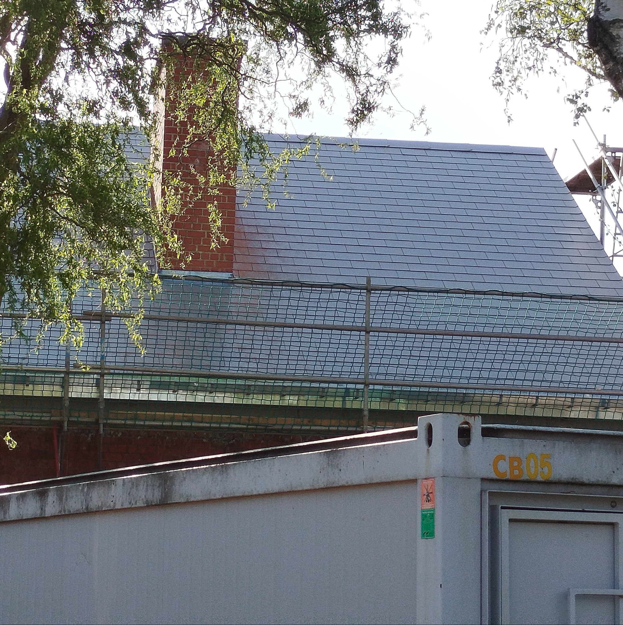 Szary dach z ceglanym kominem w tle, zabezpieczony siatką ochronną. Widoczne rusztowanie i fragment kontenera z oznaczeniem 'CB05'.