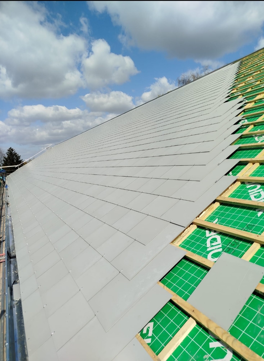 Układanie szarej dachówki na konstrukcji dachu z zieloną izolacją. Widok z góry, fragment dachu pokryty, fragment w trakcie krycia, błękitne niebo w tle.