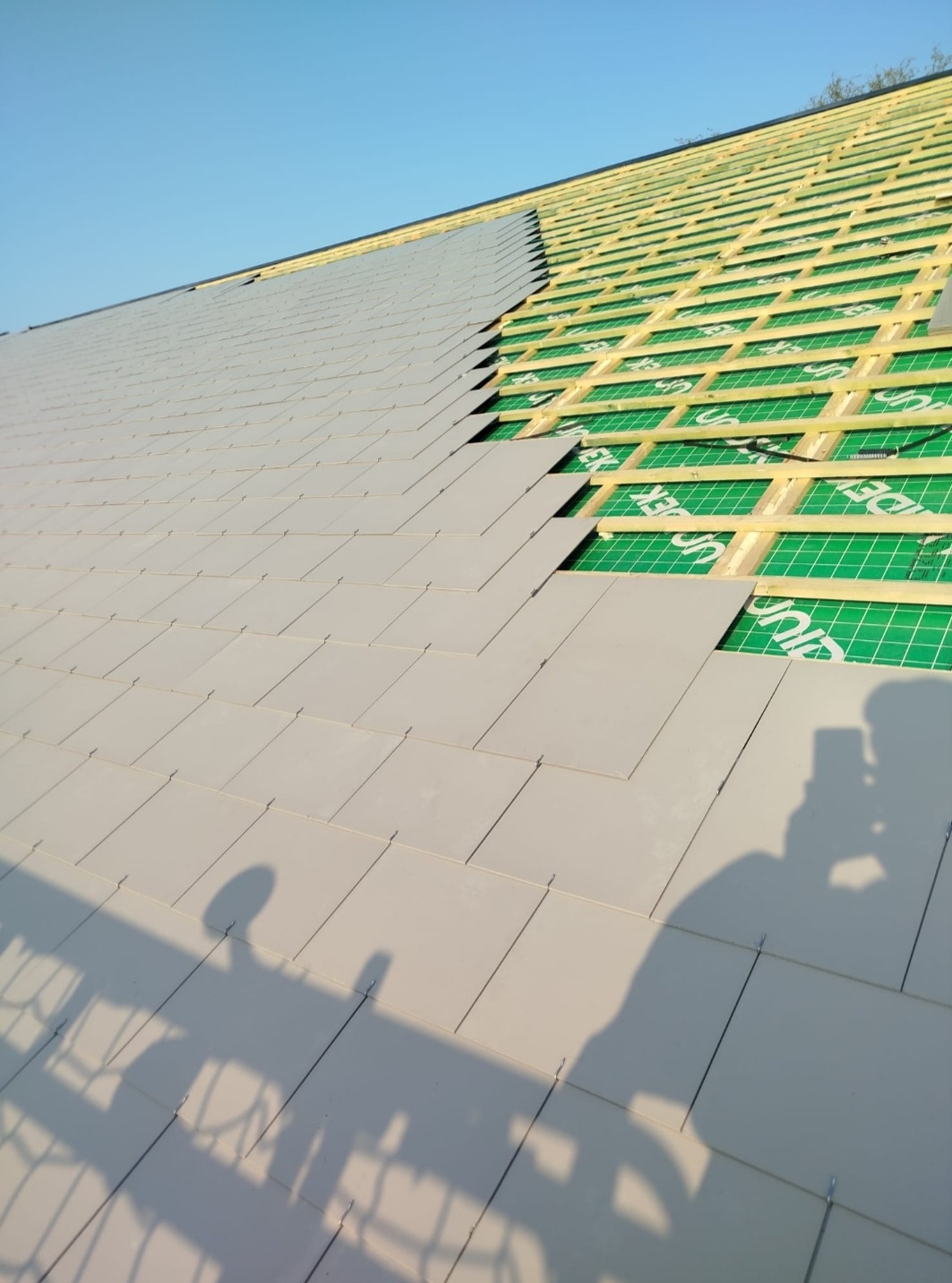 Układanie szarej dachówki na dachu z widoczną drewnianą konstrukcją i zieloną membraną. Cień osoby z telefonem w dolnej części zdjęcia.