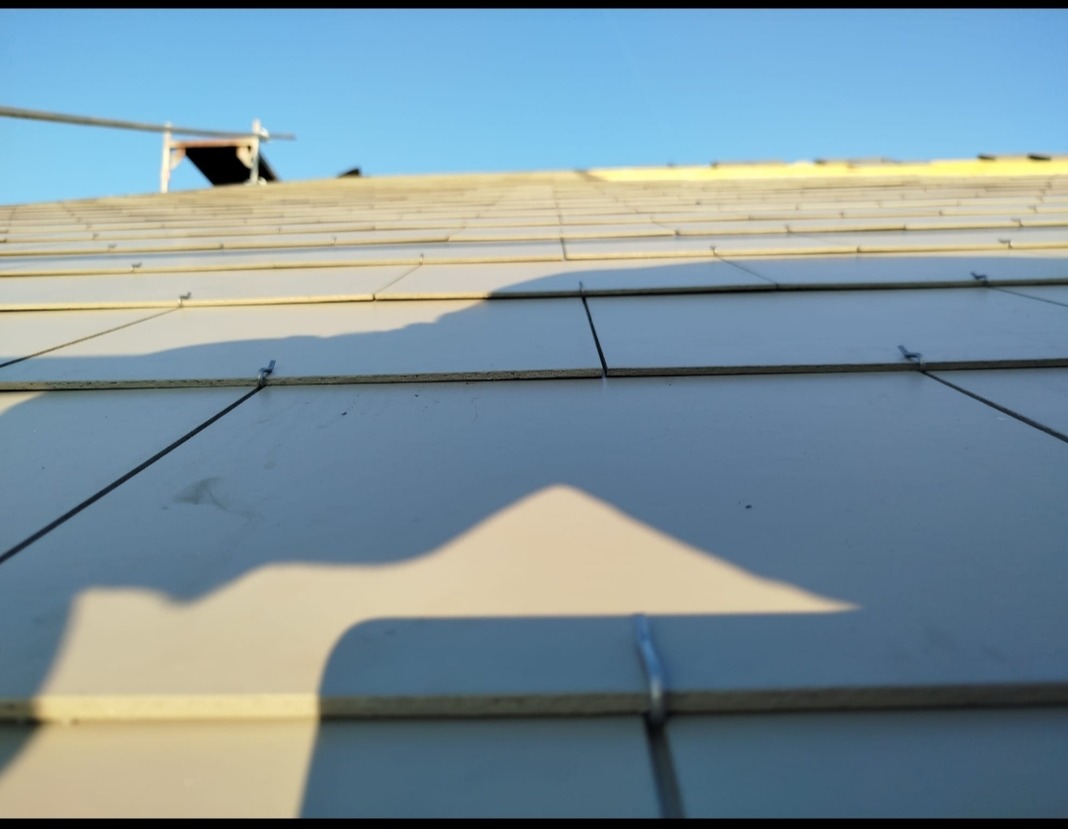Nowy dach z szarej dachówki ceramicznej, mocowany metalowymi klamrami. Widoczne fragmenty rusztowania i cień. Słoneczny dzień, błękitne niebo w tle.