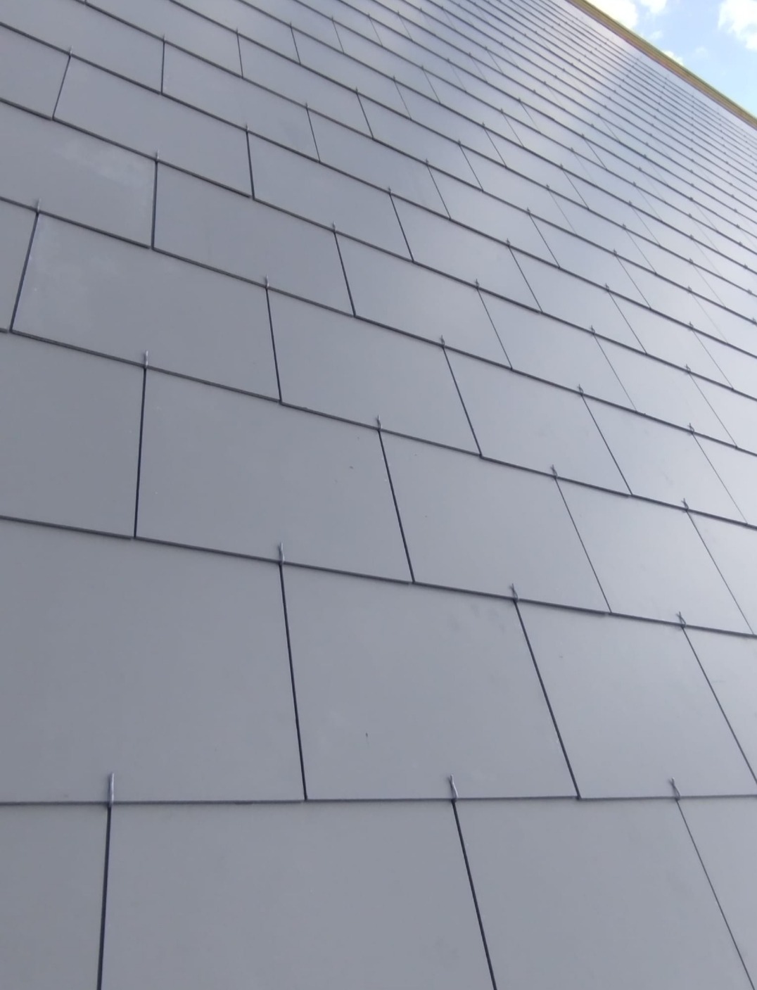 Szare, prostokątne płytki dachowe ułożone ukośnie na budynku, widoczne mocowania, fragment nieba w górnym rogu. Nowoczesne pokrycie dachu.