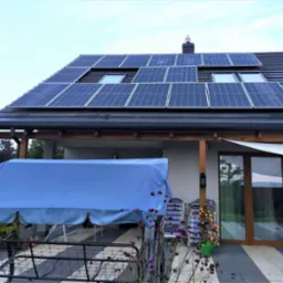 Panele słoneczne na dachu domu jednorodzinnego z oknami dachowymi, widok z poziomu tarasu z huśtawką ogrodową pod niebieskim pokrowcem.