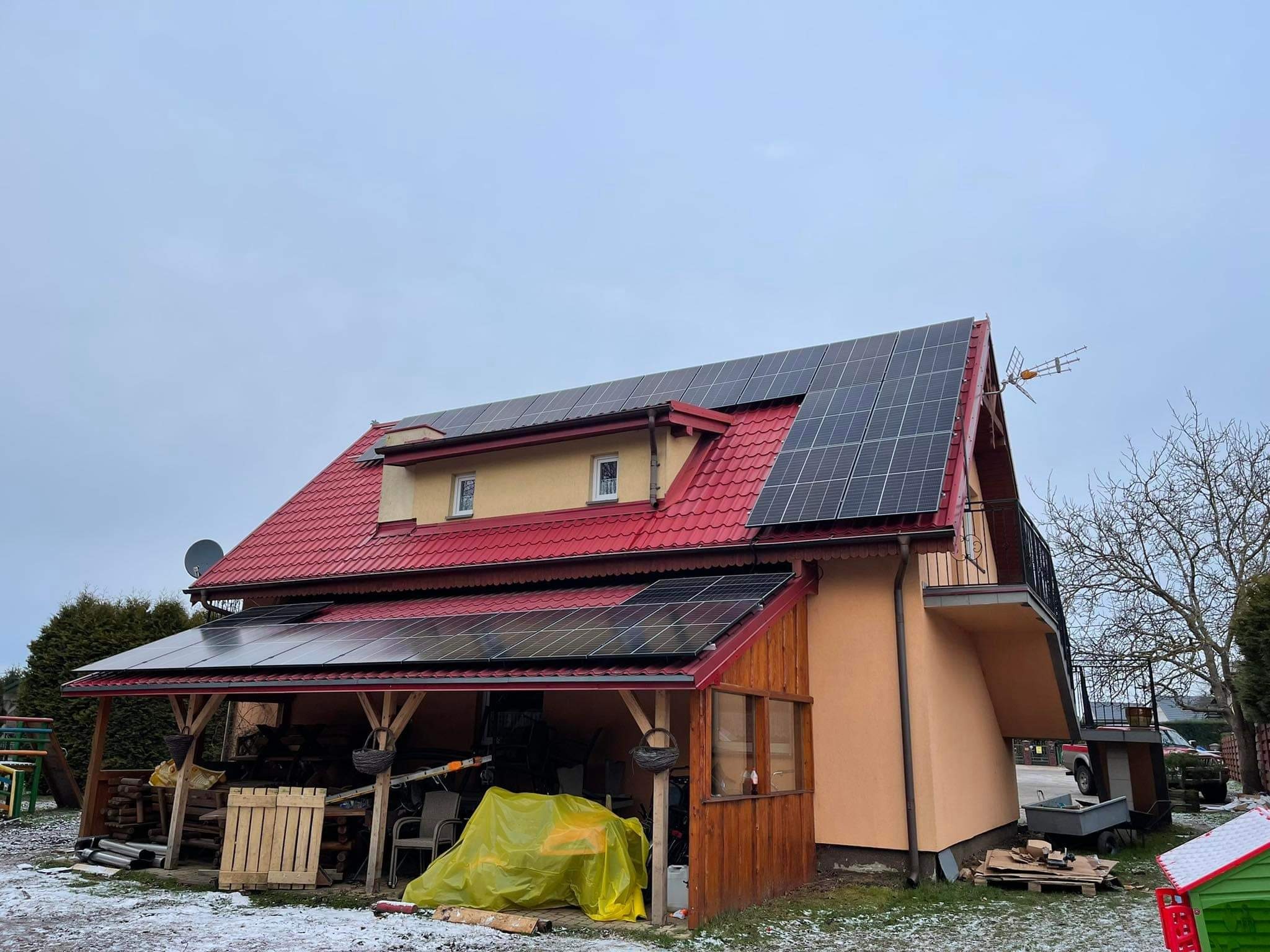 Dom jednorodzinny z czerwoną dachówką i panelami słonecznymi na dachu oraz nadbudowanym zadaszeniem, widoczne akcesoria ogrodowe i lekki śnieg na ziemi.