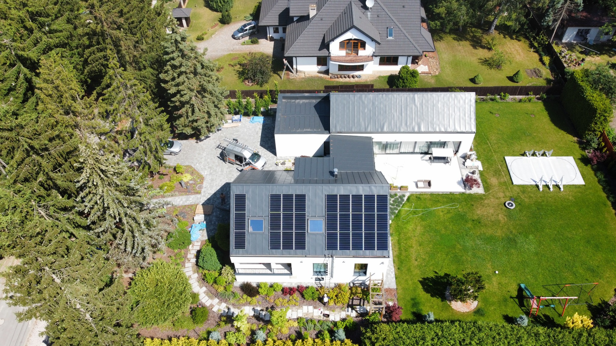 Widok z góry na dom z panelami słonecznymi na dachu, obok zaparkowany samochód z drabiną, nowoczesny ogród z basenem i placem zabaw.