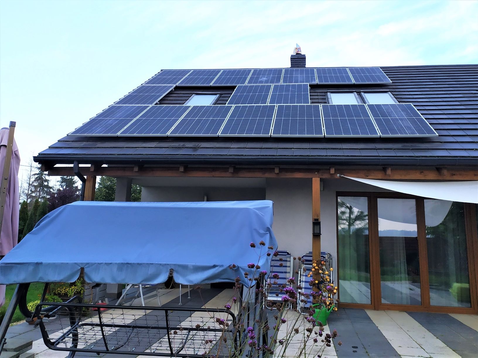 Panele słoneczne na dachu domu jednorodzinnego z oknami dachowymi, widok z poziomu tarasu z huśtawką ogrodową pod niebieskim pokrowcem.