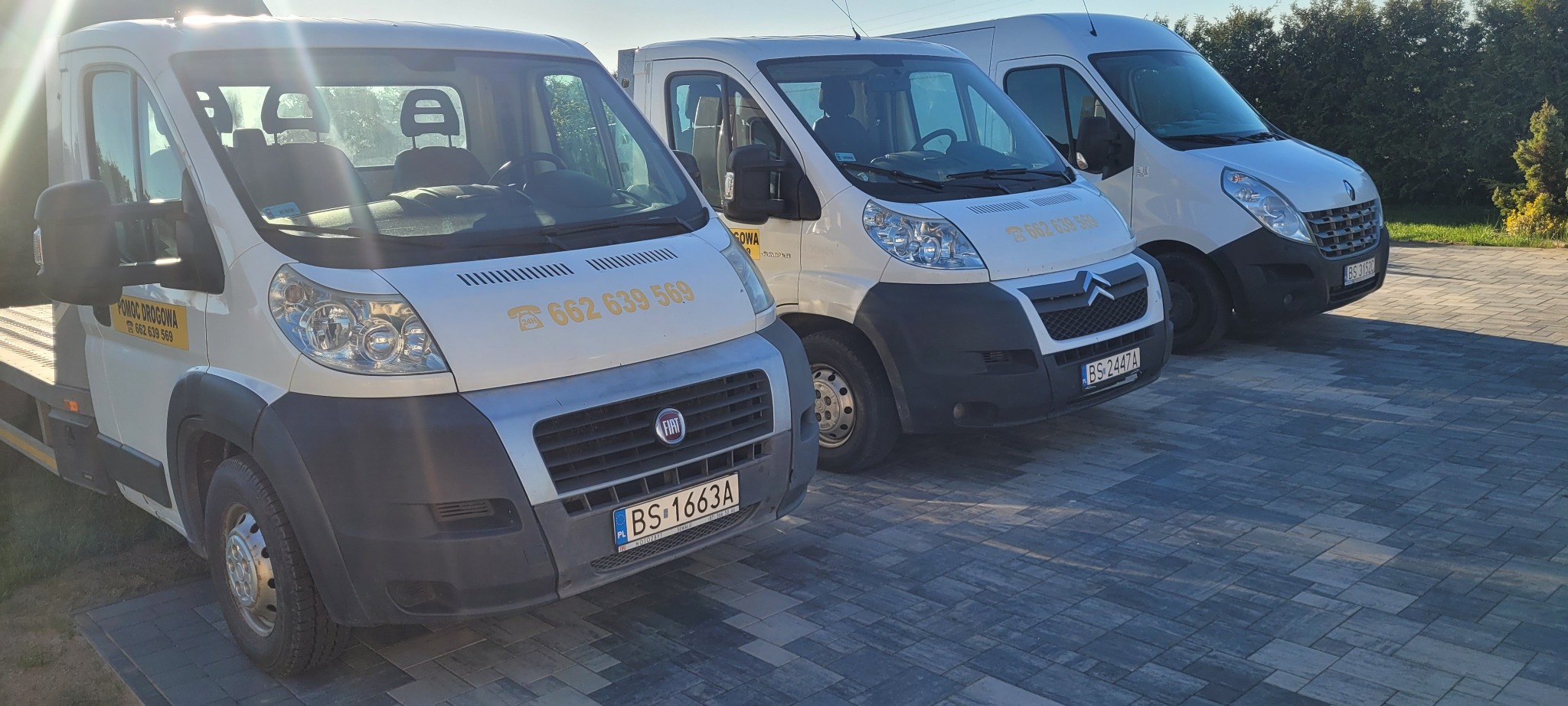 Trzy białe busy różnych marek (Fiat, Citroen, Renault) zaparkowane obok siebie na kostce brukowej, z numerami telefonów na karoseriach, sugerujące usługi transportowe lub pomoc drogową.