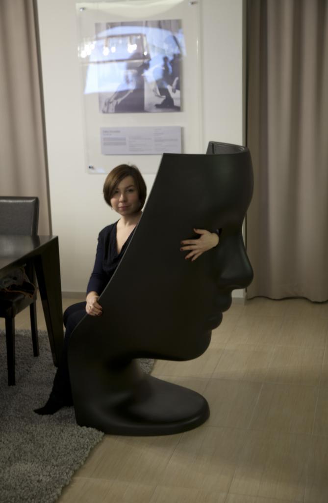 Kobieta siedzi za czarnym, designerskim krzesłem w kształcie profilu twarzy, z dłonią wystającą przez otwór na oko, w tle minimalistyczne wnętrze z zasłonami i obrazem.