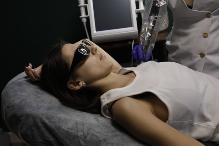 Kobieta w okularach ochronnych leży na kozetce, w trakcie zabiegu z użyciem urządzenia z osłoną, wykonywanego przez osobę w rękawiczkach i fartuchu.