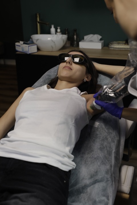 Klientka w okularach ochronnych leży na leżance podczas zabiegu depilacji laserowej pod pachami, wykonywanego przez osobę w niebieskich rękawiczkach.