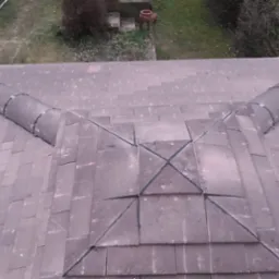 Widok z góry na dach pokryty szarą dachówką, z widocznym kominem i elementami zabezpieczającymi przed śniegiem, oraz drabina oparta o budynek.