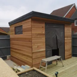 Nowoczesny drewniany domek ogrodowy z płaskim dachem, częściowo obłożony membraną, z drewnianym tarasem i drabinką w trakcie budowy.