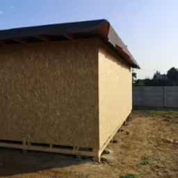 Szkieletowa konstrukcja budynku gospodarczego obita płytą OSB, z widocznym dachem pokrytym papą, osadzona na drewnianych legarach, na tle betonowego ogrodzenia i zieleni.
