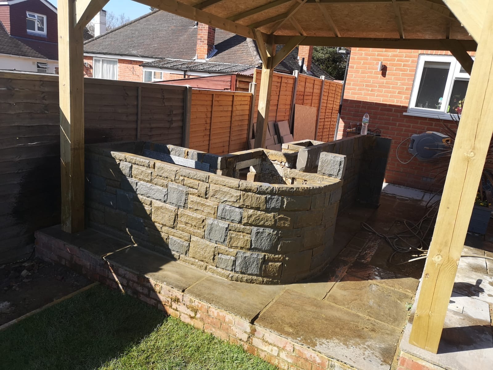 Budowa murowanego grilla z kamienia w ogrodzie, widoczna konstrukcja zadaszenia i ogrodzenie w tle, świeżo ułożona kostka brukowa.