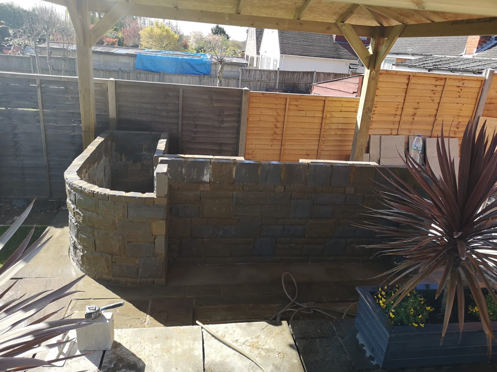 Murowana konstrukcja z kamienia w ogrodzie, otoczona drewnianym ogrodzeniem, widok zadaszenia i rośliny w donicach