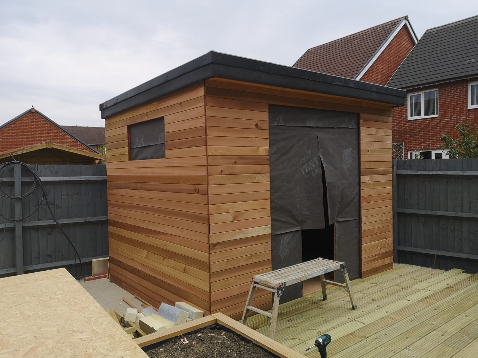 Nowoczesny drewniany domek ogrodowy z płaskim dachem, częściowo obłożony membraną, z drewnianym tarasem i drabinką w trakcie budowy.