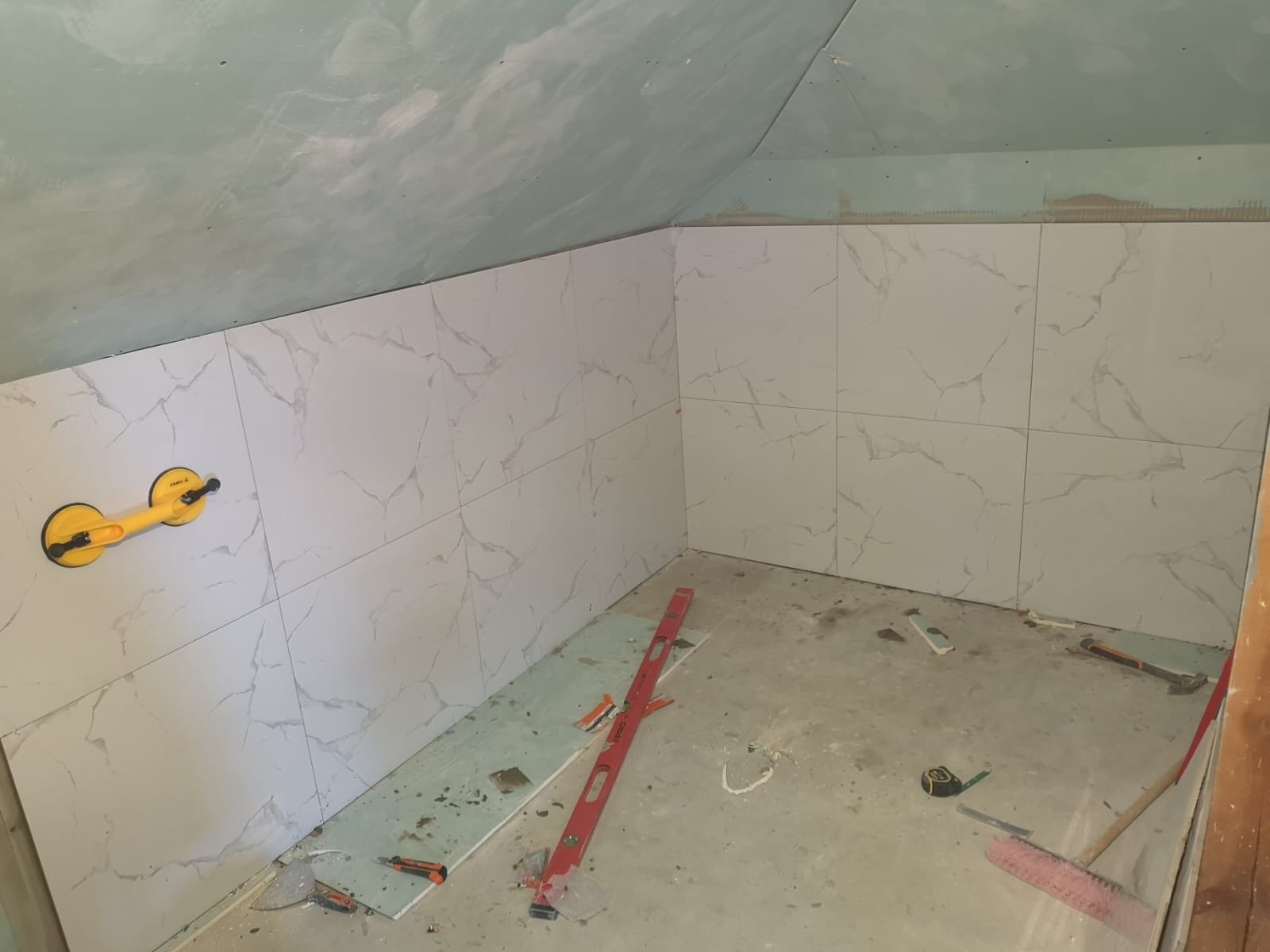 Ściana łazienki w trakcie układania białych płytek z marmurkowym wzorem, widoczna przyssawka do płytek, poziomica, fragment sufitu ze śladami szpachli i narzędzia na podłodze.