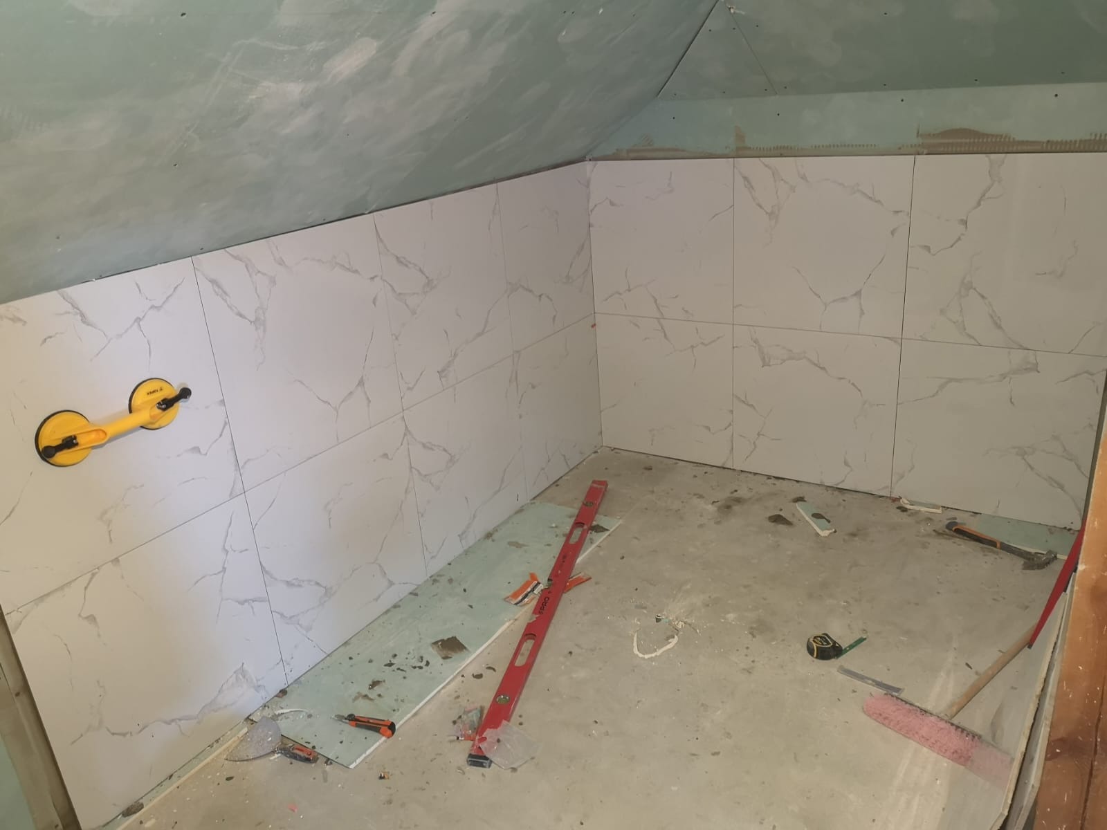 Montaż płytek imitujących marmur w łazience na poddaszu, widoczna przyssawka do płytek, poziomica i narzędzia na podłodze.
