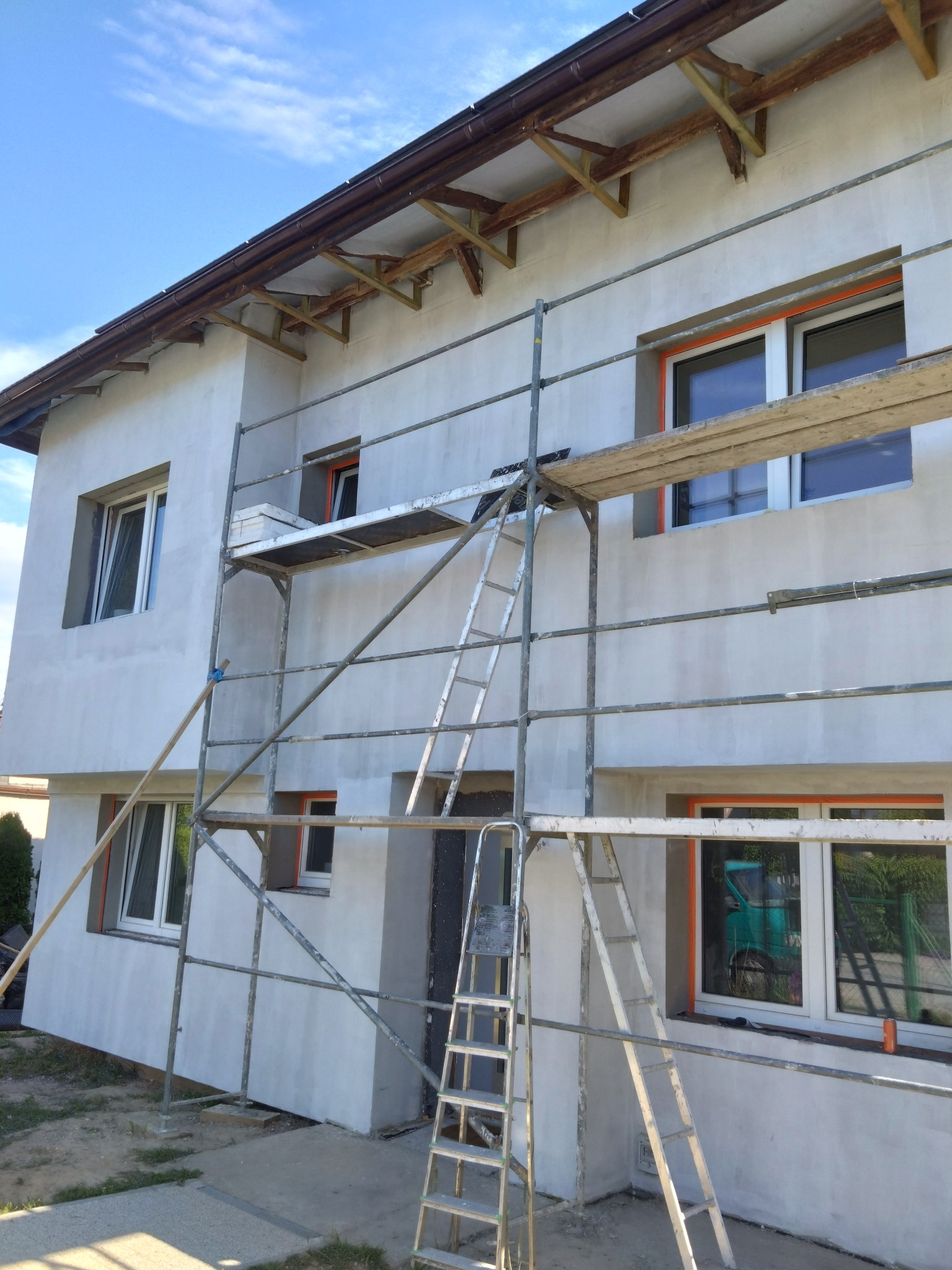 Dwupiętrowy dom w trakcie wykończenia elewacji, widoczne rusztowanie, surowa warstwa tynku na ścianach i zabezpieczone okna pomarańczową folią.