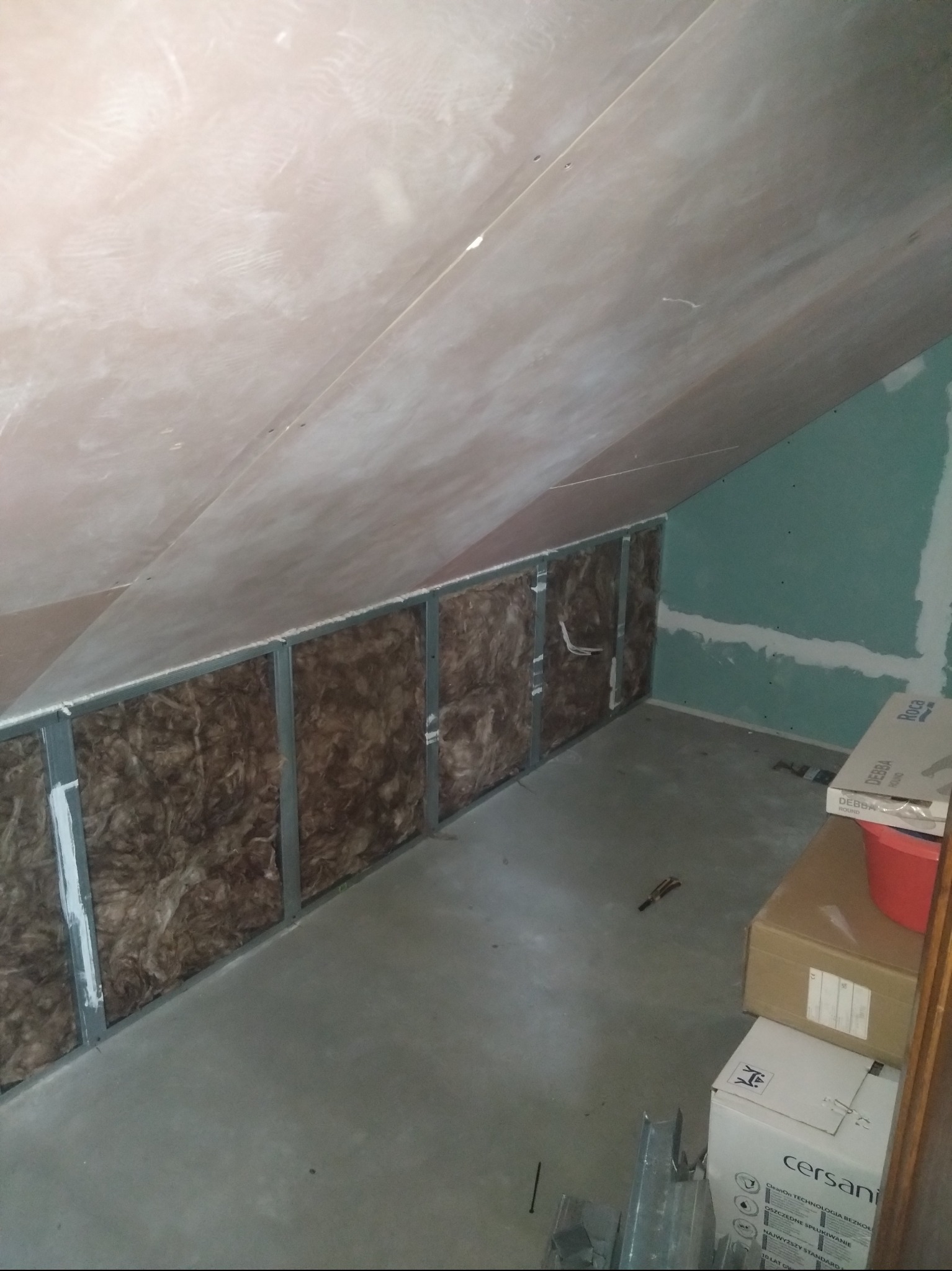 Poddasze w trakcie adaptacji: metalowy stelaż wypełniony wełną mineralną, płyty gipsowo-kartonowe na ścianie, surowy beton na podłodze, kartony z ceramiką sanitarną w rogu.