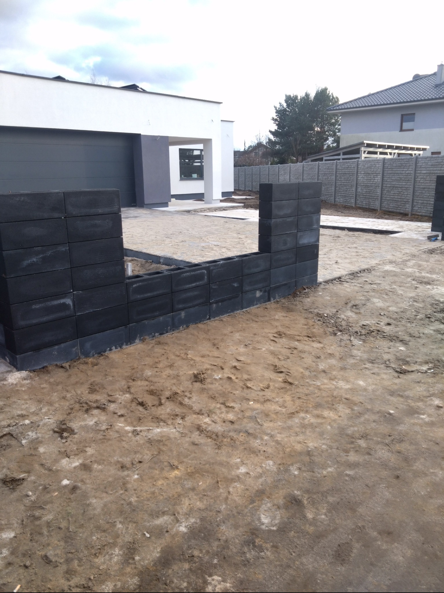 Budowa ogrodzenia z szarych, betonowych bloczków przy nowoczesnym domu z garażem, widoczne ślady prac ziemnych i fragment gotowego ogrodzenia panelowego w tle.