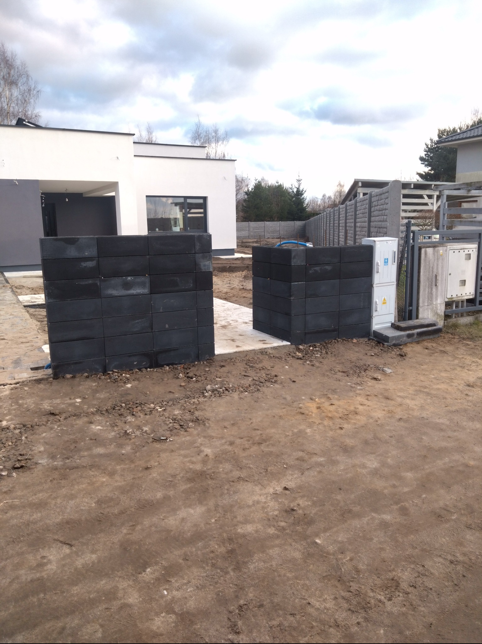 Wzniesione słupki z szarej kostki betonowej na fundamencie, prawdopodobnie pod bramę wjazdową, na tle nowoczesnego domu z płaskim dachem i ogrodzenia z betonowych prefabrykatów.