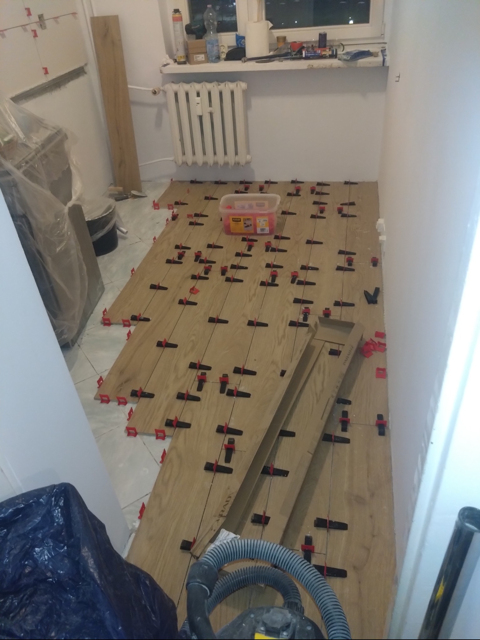 Układanie paneli podłogowych imitujących drewno z użyciem systemu poziomowania płytek, widoczny odkurzacz i opakowanie po systemie poziomowania.